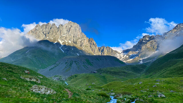 Morning clouds gather around Chaukhi Mountain above green slopes and lingering snow in Georgia, illustrating alpine weather