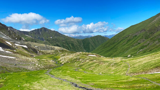 An alpine valley unfolds along the Roshka to Juta route in Georgia, where a narrow trail, melting snowfields, rocky slopes