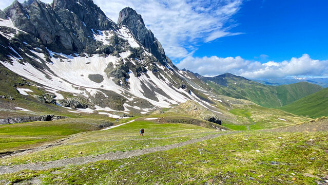 Backpacker hikes from Roshka toward Juta across wide alpine valley in Georgia, passing snow-covered peaks and remote terrain