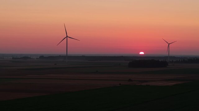 A beautiful view of a wind farm during golden hour, with the sun shining in a bright orange sky and multiple wind turbines silhouetted against the field.