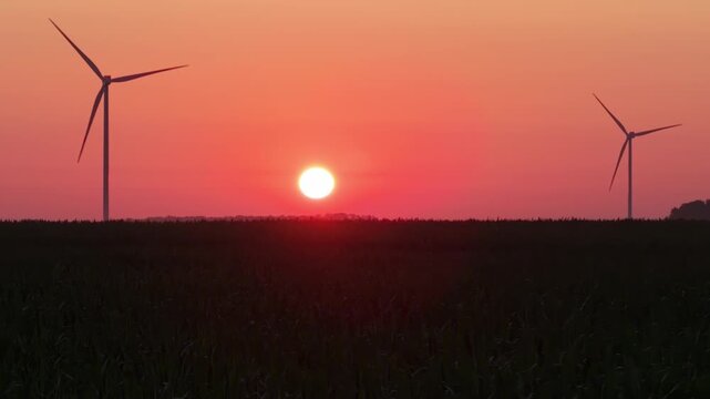 A beautiful view of a wind farm during golden hour, with the sun shining in a bright orange sky and multiple wind turbines silhouetted against the field.