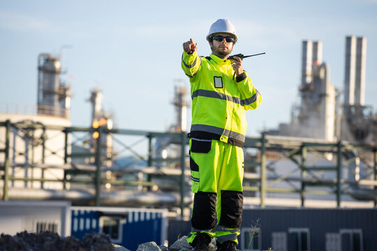 industrial engineer pointing forward holding walkie talkie at oil refinery plant, Worker man in high visibility jacket, Site inspector with radio pointing directions at petrochemical facility