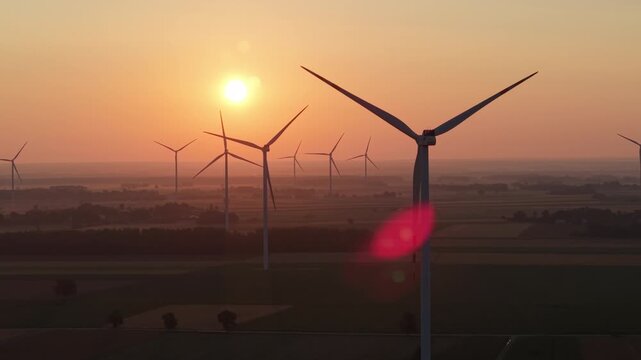 A beautiful view of a wind farm during golden hour, with the sun shining in a bright orange sky and multiple wind turbines silhouetted against the field.