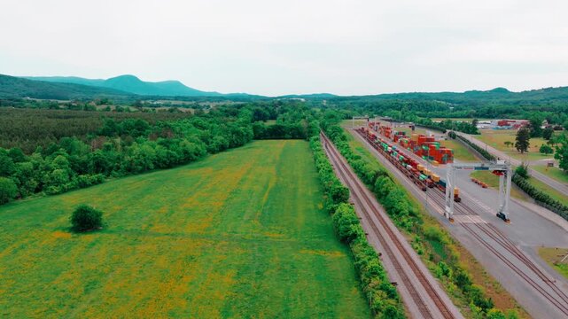 Freight locomotive hauling intermodal cars along twin tracks through green Appalachian countryside toward a container terminal with cranes.