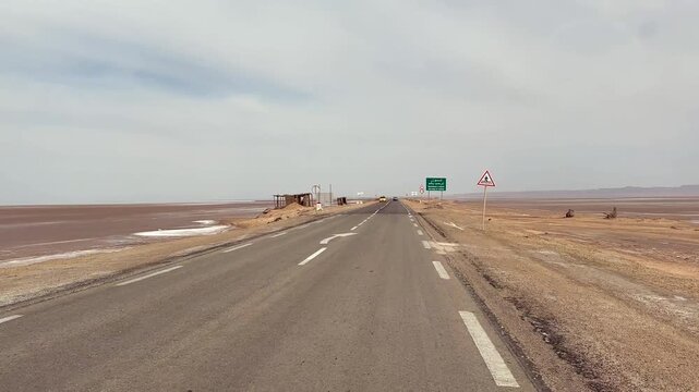 Desert highway runs across the dry Chott el Djerid salt flat with roadside signs reading Au Revoir and Good Bye under a pale daytime sky. Tunisia.