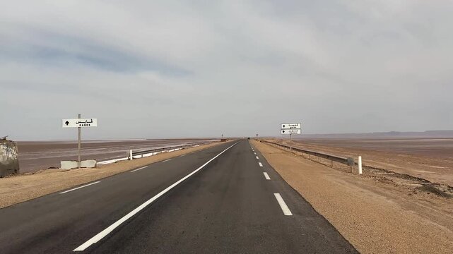 Desert highway runs across the dry Chott el Djerid salt flat under a pale daytime sky, Tunisia.