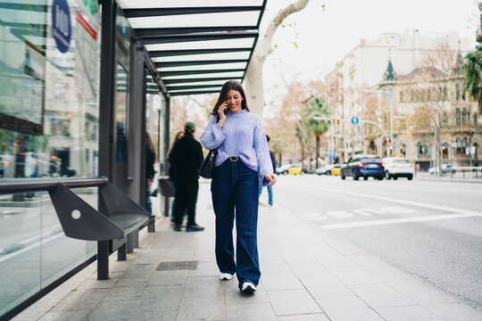 Happy young woman walking at bus stop and talking on smartphone, mobile conversation, 5G connection, roaming service, urban background, modern lifestyle, digital communication, city street