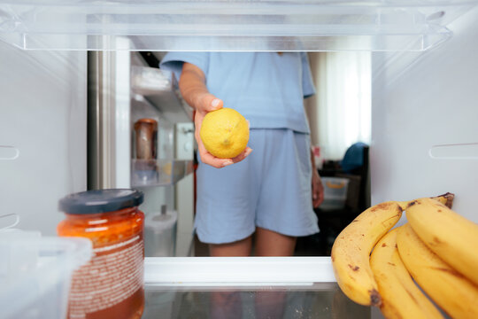 Woman Reaching For Fresh Lemon Inside Open Refrigerator. Girl grabbing yellow citrus fruit from shelf viewed from inside kitchen fridge