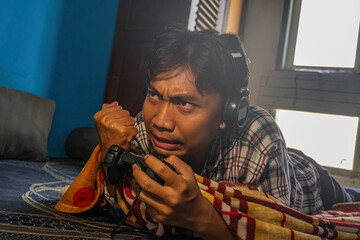 An asian man lying on bed with headphones, playing video games with a gamepad controller and showing intense emotion. Casual gaming and youth indoor lifestyle concept. © Anzz Stock