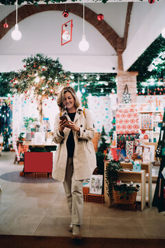 Smiling woman using smartphone while walking in festive holiday shop, middle aged female browsing mobile application for gift shopping, authentic portrait of digital inclusion and positive mood.