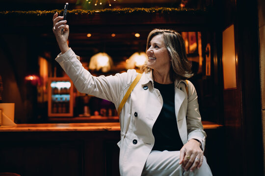 Smiling woman taking selfie on smartphone at cafe, happy middle aged lady vlogging using mobile phone camera, authentic portrait of digital nomad influencer lifestyle and tech connectivity.