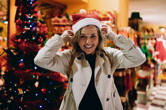 Smiling woman posing with Santa hat near decorated Christmas tree, joyful female traveler enjoying winter holiday atmosphere, authentic portrait of festive celebration and positive attitude.