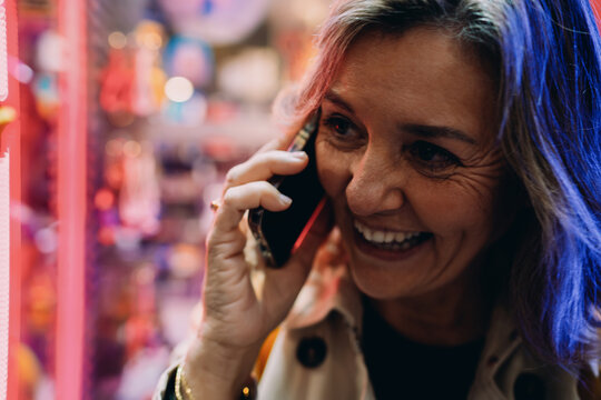 Smiling adult female having emotional phone conversation in urban setting, joyful woman using smartphone for mobile call, authentic representation of social connection and modern technology.