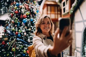 Portrait of smiling mature woman looking at smartphone screen to take selfie, happy female posing with mobile phone near Christmas tree, digital communication, authentic representation, tech © BullRun