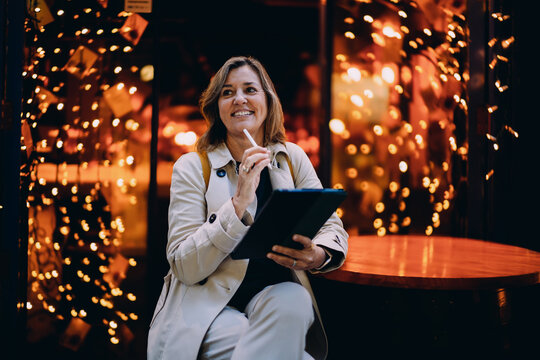 Inspired woman looking away while working on tablet at night cafe, female freelancer dreaming of success holding stylus pen, ambition, creative vision, digital nomad, copy space, bokeh lights