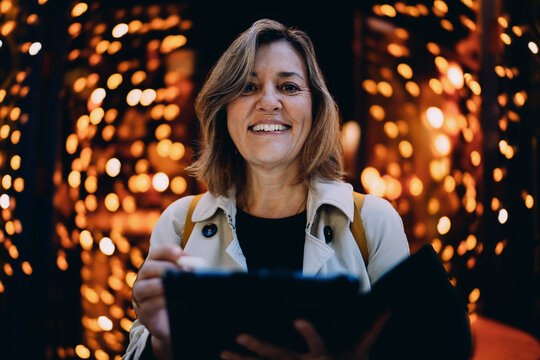 Cheerful middle aged woman looking at camera holding tablet and stylus pen in night city, portrait of professional female freelancer using modern gadget outdoors, authentic representation