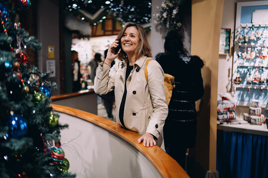 Positive mature woman using smartphone for calling friends in festive indoor market, happy female during mobile conversation near Christmas decorations, 5G connection, lifestyle photography