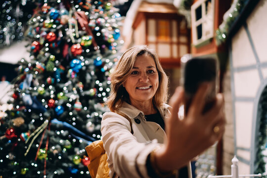 Portrait of smiling mature woman looking at smartphone screen to take selfie, happy female posing with mobile phone near Christmas tree, digital communication, authentic representation, tech
