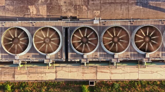 Above view shows cooling towers in a row at a facility. The sun shines down and creates shadow patterns on the ground. Green grass is visible next to the industrial area.
