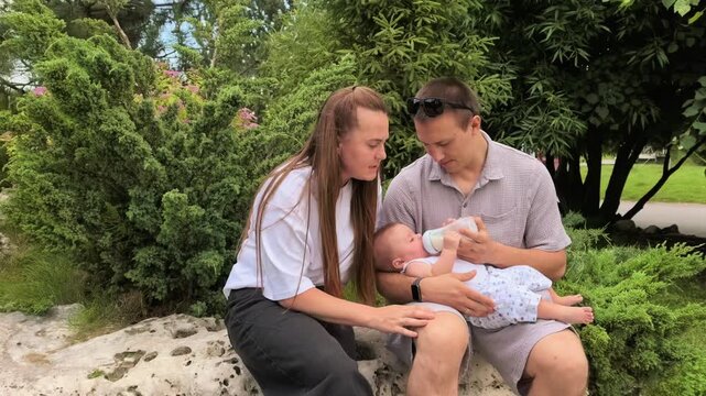 Father bottle feeds an infant while the mother watches outdoors in a park, loving family care, parenthood, baby nutrition, tender bonding.