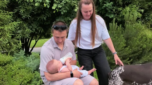 Father bottle feeds an infant while sitting with a woman and dog in a park, family outing, baby care, pet companionship, outdoor lifestyle.