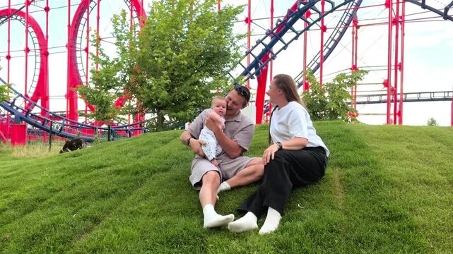 Father lifts an infant while the mother watches at an amusement park, joyful family bonding, playful parenthood, outdoor leisure, childhood fun.
