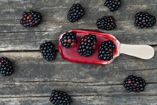 A popsicle rests on a rustic wooden board. Blackberries are scattered on top of the popsicle and around it. It's summer and hot, and the popsicle is starting to melt.