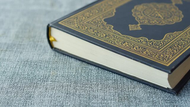 Close up video of a sacred Islamic book, Quran, with ornate gold lettering on a dark blue cover, placed on a textured grey fabric surface