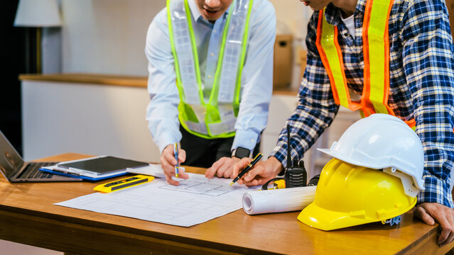 Two construction workers planning a project over blueprints at a desk with laptop, tools, and safety helmets in an office setting