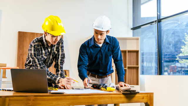 Two construction workers in hard hats reviewing plans at a worktable with laptop and tools in a bright modern office