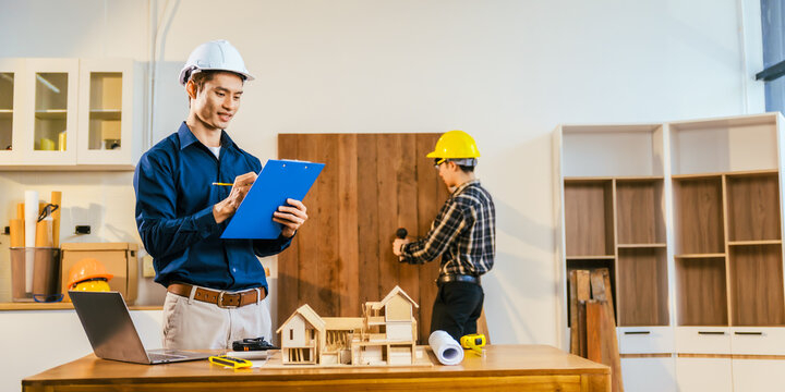 Two construction workers planning a home renovation in a workshop, one writing on a clipboard while another drills wood next to a house model and laptop