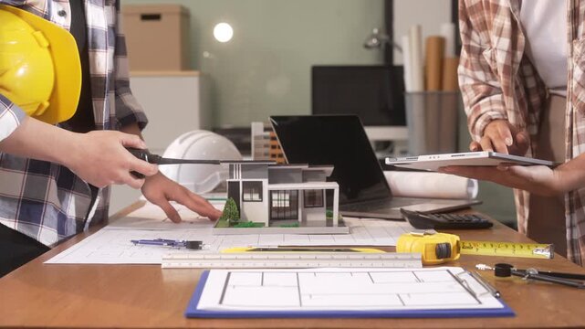 Two people reviewing a house model, blueprint and tools at a desk during an architecture meeting, with laptop, hard hat and plan papers.