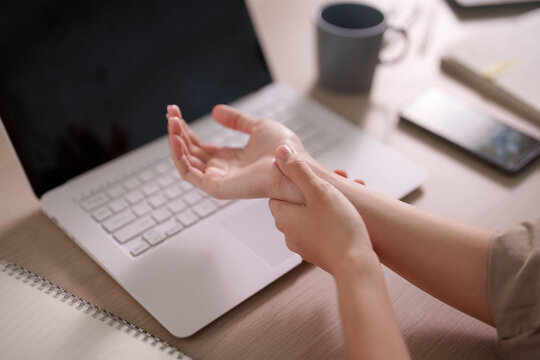 Asian woman massaging painful wrist experiencing carpal tunnel syndrome while working on laptop at home office