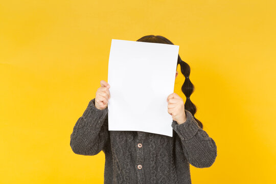 Playful little girl hiding her face behind a blank white sheet of paper, standing against a bright yellow studio background.
