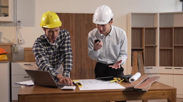 Two construction workers reviewing blueprints and laptop plans in a home renovation workspace with tools and wood samples