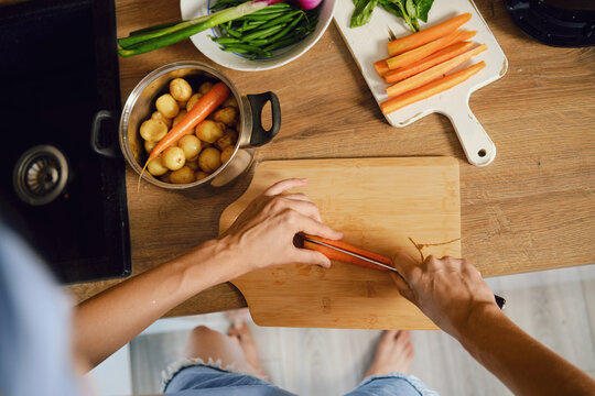 Top view of hands slicing fresh carrot lengthwise on wooden chopping board in kitchen