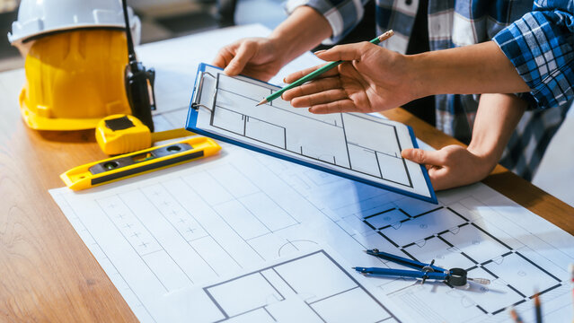 Close up of architects discussing a house floor plan on a desk with blueprints, helmet, level and drawing tools in a bright office.
