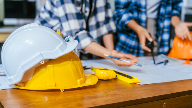 White and yellow safety helmets on a wooden table with workers reviewing blueprints in the background, showing construction planning, teamwork and workplace safety.