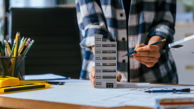 Architect holding a small building model over blueprints on a desk in a modern office, planning a construction project with pencils and tools nearby.