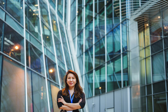 Confident asian business woman with arms crossed, smiling in front of a glass skyscraper