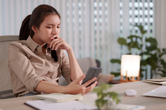 Worried Asian businesswoman biting nail while reading smartphone message in modern home office workspace