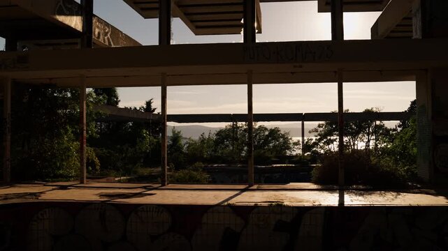 Sunset shines through the abandoned Haludovo hotel on Krk Island, revealing graffiti, concrete columns, overgrown vegetation, and the Adriatic Sea beyond.