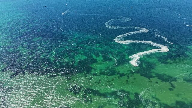 Racers navigate jet skis at high speed in a body of clear blue water during daytime. White trails mark their paths across the surface. People enjoy the activity on a sunny day.