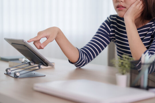 Young Asian woman looking stressed and thoughtful while using tablet on stand at home workspace with copy space