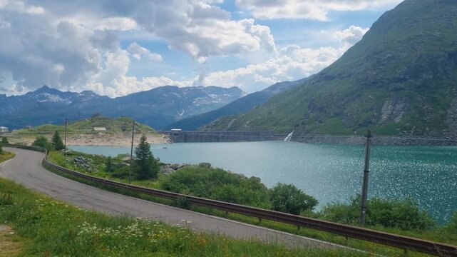 Scenic high-altitude view of Lago di Montespluga near Spl&uuml;gen Pass, Italy, showcasing a pristine alpine reservoir framed by rugged mountain peaks and a winding alpine road under a partly cloudy sky