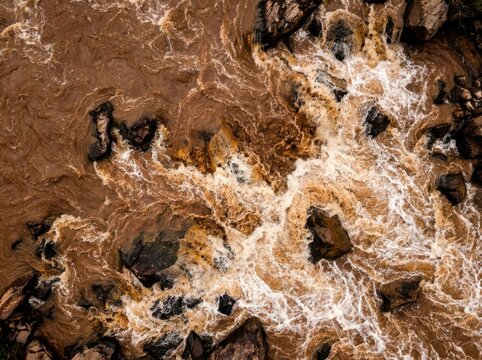 Turbulent brown river water with white foam swirling around dark rocks aerial view powerful natural current