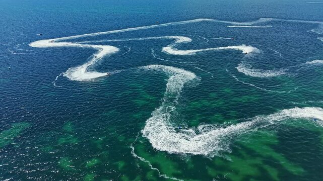 Riders on jet skis race across clear water, creating splashes and trails. The competition is intense as they navigate curves and speed along the course. The scene takes place on a sunny day.