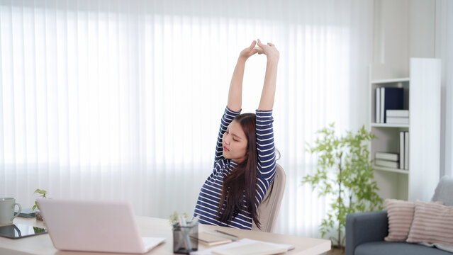 Young Asian woman stretching arms overhead during work break at home office desk with copy space