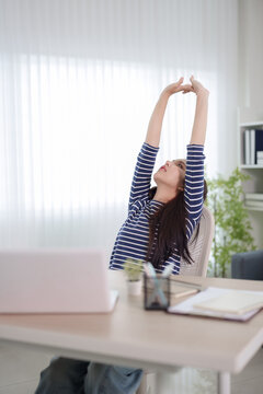 Young Asian woman stretching arms overhead during work break in bright home office with copy space