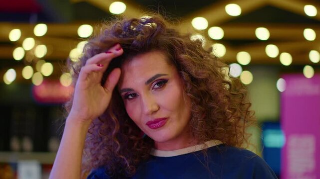 Woman adjusts her voluminous curls. Her gentle smile faces the camera. Warm bokeh lights fill the background. Her blue top enhances casual charm. Ideal for beauty and fashion promotions